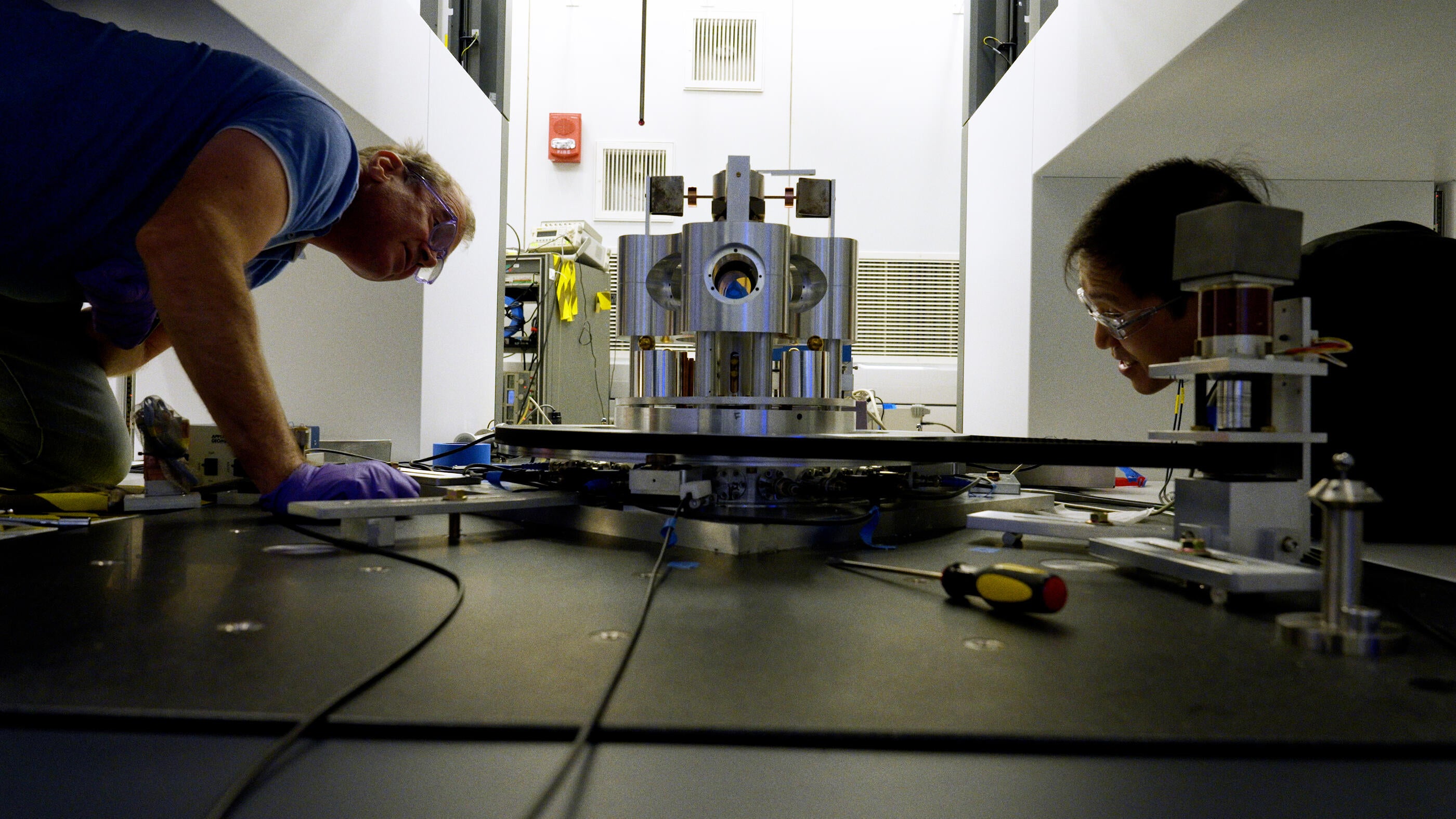 Two men wearing goggles leaning over a table looking at a torsion balance machine